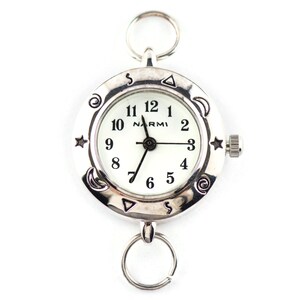 May include: A silver-toned pocket watch with a white face and black numerals. The brand name NARMI is printed on the dial. The bezel features engraved symbols including stars, moons, and swirls. It has a circular ring at the top and bottom.