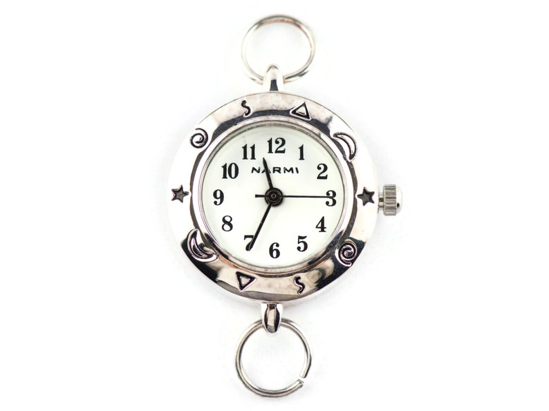 May include: A silver-toned pocket watch with a white face and black numerals. The brand name NARMI is printed on the dial. The bezel features engraved symbols including stars, moons, and swirls. It has a circular ring at the top and bottom.