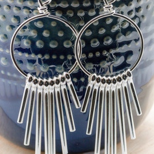 Puede incluir: Pendientes de aro de plata con una franja de barras de plata colgantes. Los pendientes cuelgan de una superficie con estampado azul y blanco.