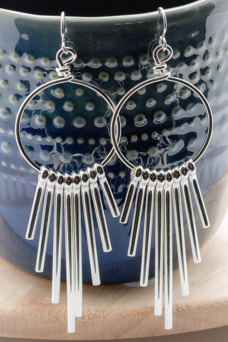Puede incluir: Pendientes de aro de plata con una franja de barras de plata colgantes. Los pendientes cuelgan de una superficie con estampado azul y blanco.