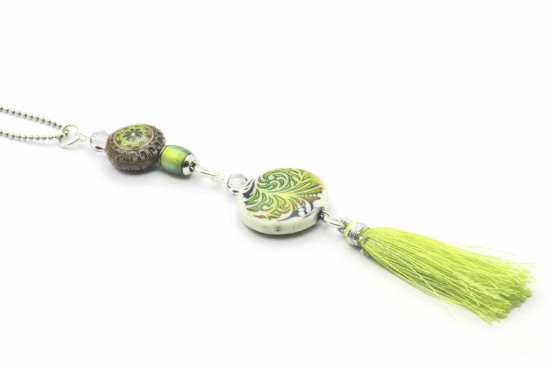 May include: A green and brown beaded necklace with a green tassel. The necklace features a round ceramic pendant with a green and white floral design.