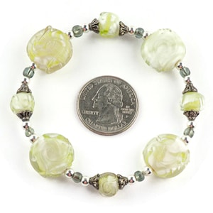Peut inclure: Un bracelet en verre vert avec des motifs de fleurs grav&eacute;s et des accents argent&eacute;s. Le bracelet est &agrave; peu pr&egrave;s de la taille d'une pi&egrave;ce de 25 centimes.