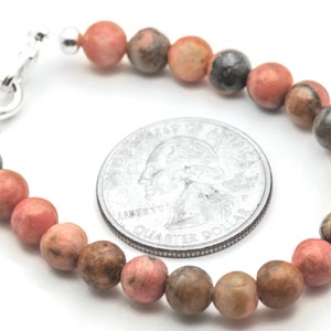 K&ouml;nnte beinhalten: Ein Armband aus rosafarbenen, braunen und schwarzen Edelsteinen mit einem silbernen Verschluss. Das Armband ist etwa so gro&szlig; wie eine 25-Cent-M&uuml;nze.