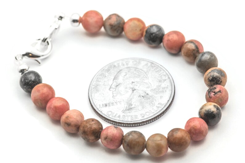 K&ouml;nnte beinhalten: Ein Armband aus rosafarbenen, braunen und schwarzen Edelsteinen mit einem silbernen Verschluss. Das Armband ist etwa so gro&szlig; wie eine 25-Cent-M&uuml;nze.