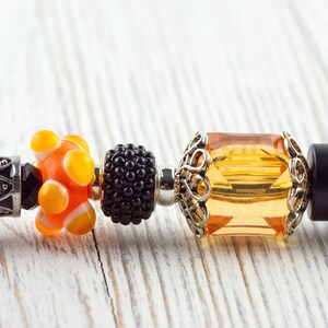 May include: Close-up of a bracelet with decorative beads. The beads include a black and silver bead, an orange and yellow bead, a black bead, and a large, square, amber-colored bead with a silver filigree setting. The bracelet is on a white wooden surface.