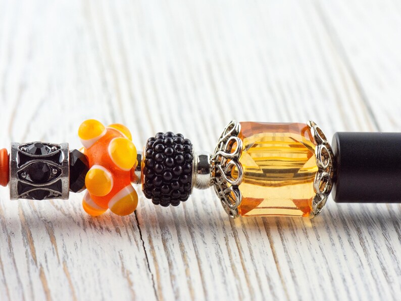 May include: Close-up of a bracelet with decorative beads. The beads include a black and silver bead, an orange and yellow bead, a black bead, and a large, square, amber-colored bead with a silver filigree setting. The bracelet is on a white wooden surface.