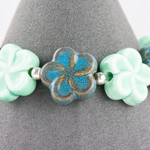 May include: A bracelet made of ceramic flower beads in shades of turquoise, blue, and green. The beads are strung on a silver cord.