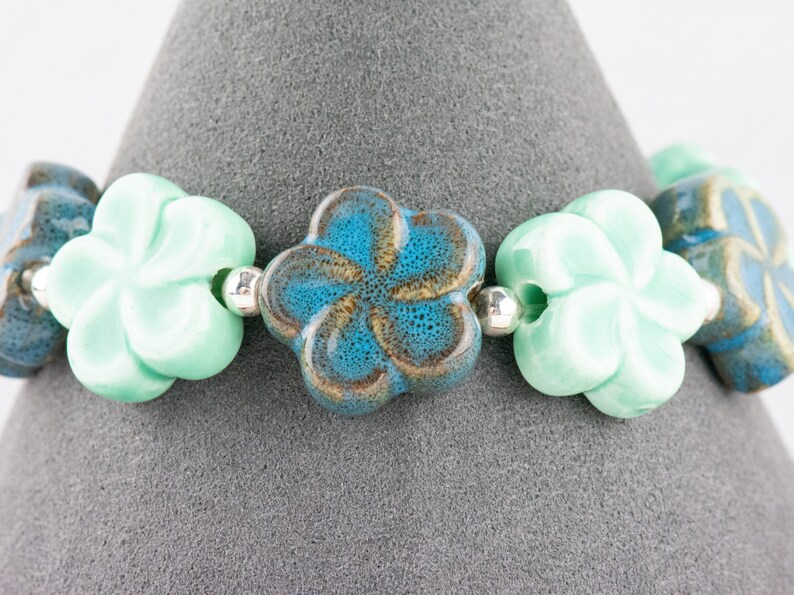 May include: A bracelet made of ceramic flower beads in shades of turquoise, blue, and green. The beads are strung on a silver cord.