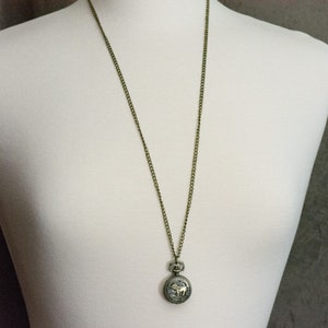 May include: A gold chain necklace with a round, silver pocket watch pendant.