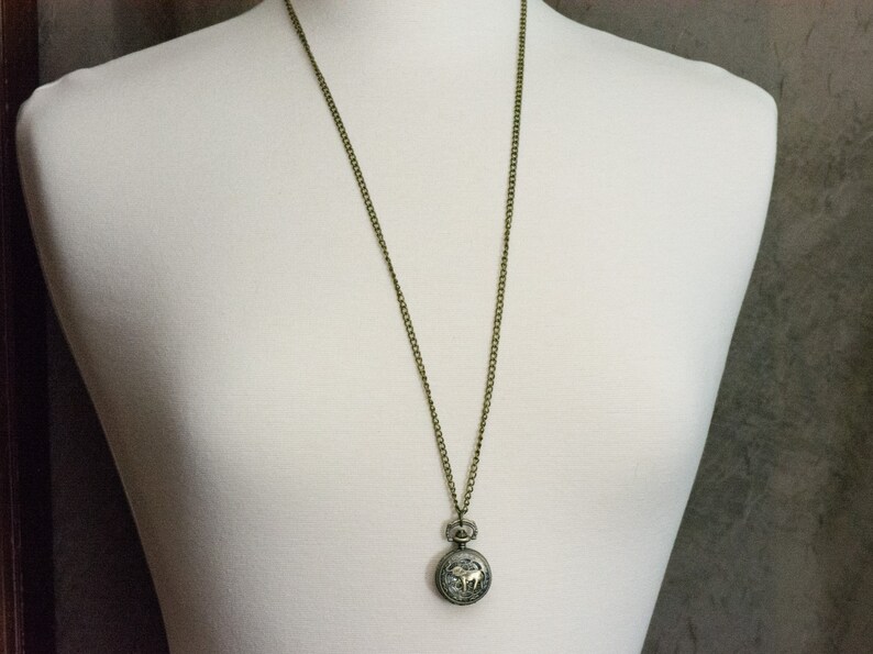 May include: A gold chain necklace with a round, silver pocket watch pendant.