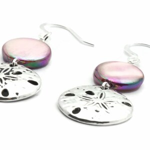 Puede incluir: Pendientes de plata con un dije de concha iridiscente rosa y morado y un dije de d&oacute;lar de arena plateado.