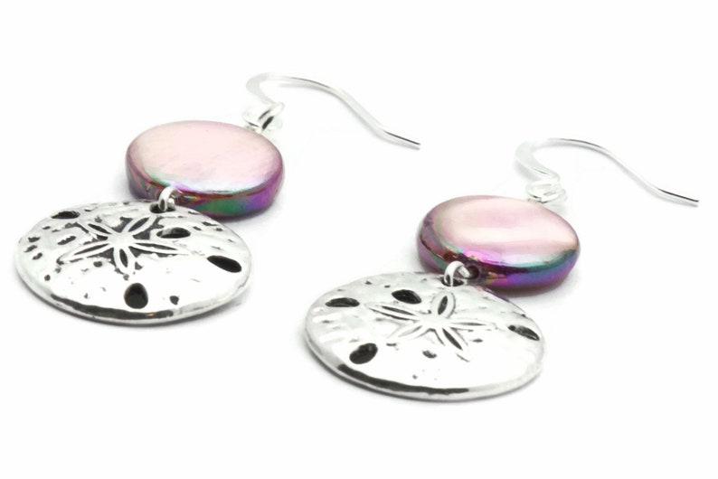 Puede incluir: Pendientes de plata con un dije de concha iridiscente rosa y morado y un dije de d&oacute;lar de arena plateado.