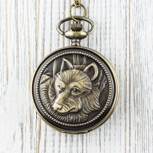 May include: A gold-toned pocket watch with a wolf head design on the face. The watch has a chain attached to it.