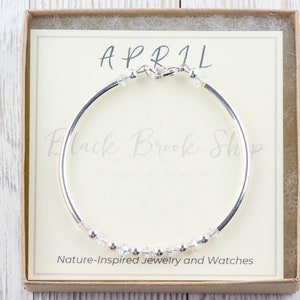 K&ouml;nnte beinhalten: Ein silbernes Armreif-Armband mit einer Reihe von klaren Kristallen und dem Wort "APRIL" auf einer wei&szlig;en Karte in einer braunen Schachtel. Die Karte tr&auml;gt au&szlig;erdem den Schriftzug "Black Brook Shop" und "Naturinspirierte Schmuckst&uuml;cke und Uhren".