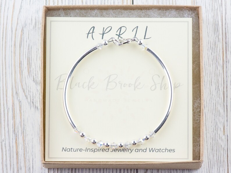 K&ouml;nnte beinhalten: Ein silbernes Armreif-Armband mit einer Reihe von klaren Kristallen und dem Wort "APRIL" auf einer wei&szlig;en Karte in einer braunen Schachtel. Die Karte tr&auml;gt au&szlig;erdem den Schriftzug "Black Brook Shop" und "Naturinspirierte Schmuckst&uuml;cke und Uhren".