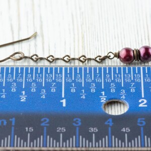 Puede incluir: Pendiente de bronce antiguo con una cadena en forma de coraz&oacute;n y dos cuentas de color p&uacute;rpura oscuro. El pendiente mide aproximadamente 5 cm de largo, medido por la regla en la imagen. El gancho del pendiente es de dise&ntilde;o simple y curvado.