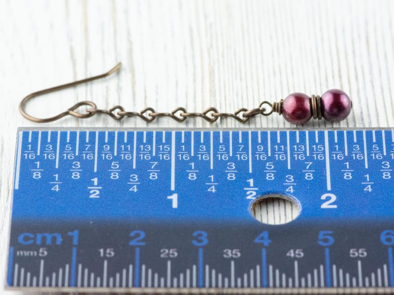 Puede incluir: Pendiente de bronce antiguo con una cadena en forma de coraz&oacute;n y dos cuentas de color p&uacute;rpura oscuro. El pendiente mide aproximadamente 5 cm de largo, medido por la regla en la imagen. El gancho del pendiente es de dise&ntilde;o simple y curvado.