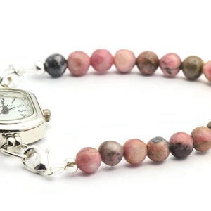 K&ouml;nnte beinhalten: Eine silberne Uhr mit quadratischem Zifferblatt und einem Armband aus rosa und braunen Perlen.