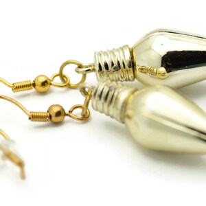 Puede incluir: Un par de pendientes de tono dorado con un dije de bombilla de luz plateada. Las bombillas son brillantes y tienen una punta puntiaguda.