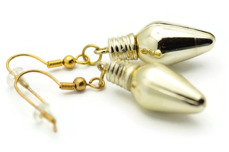 Puede incluir: Un par de pendientes de tono dorado con un dije de bombilla de luz plateada. Las bombillas son brillantes y tienen una punta puntiaguda.