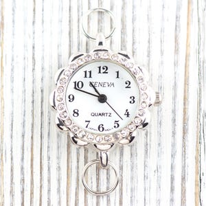 May include: A silver-toned Geneva quartz watch with a white face and a floral-shaped bezel encrusted with rhinestones. The watch has a ring at the top and bottom for attachment. The watch hands point to 10:09.
