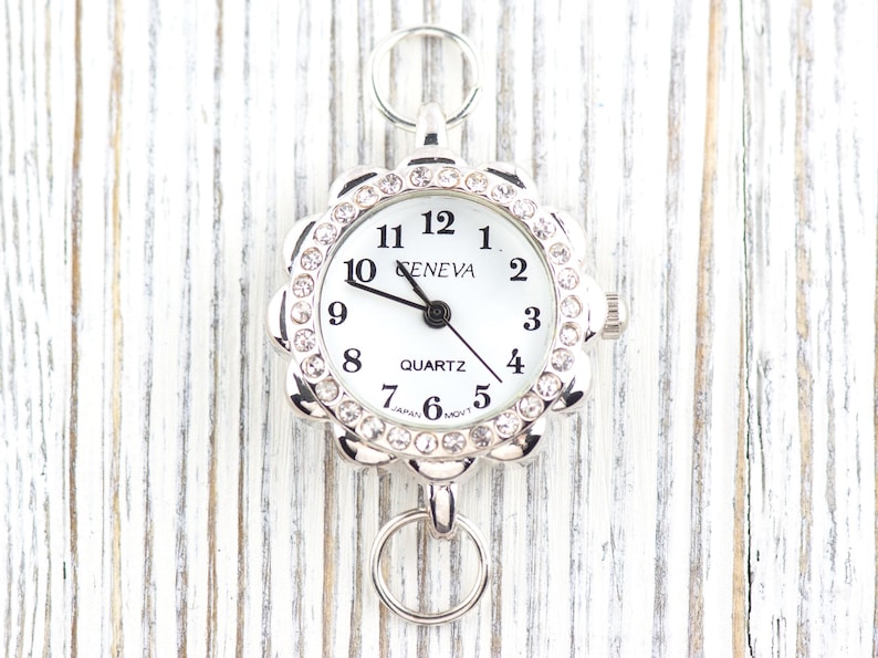 May include: A silver-toned Geneva quartz watch with a white face and a floral-shaped bezel encrusted with rhinestones. The watch has a ring at the top and bottom for attachment. The watch hands point to 10:09.