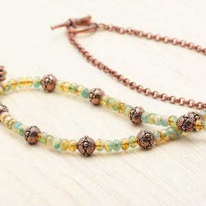 Pu&ograve; includere: Una collana artigianale con una catena color rame e un filo di perline con pietre preziose gialle e verde-blu. La collana include perline decorative in rame e un grande ciondolo dettagliato. La collana &egrave; esposta su una superficie di legno chiaro.