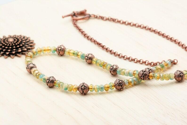 Pu&ograve; includere: Una collana artigianale con una catena color rame e un filo di perline con pietre preziose gialle e verde-blu. La collana include perline decorative in rame e un grande ciondolo dettagliato. La collana &egrave; esposta su una superficie di legno chiaro.