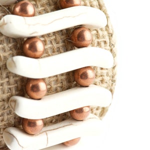 May include: A close-up of a bracelet made with white ceramic beads and copper-colored metal beads. The bracelet is wrapped around a piece of burlap fabric.