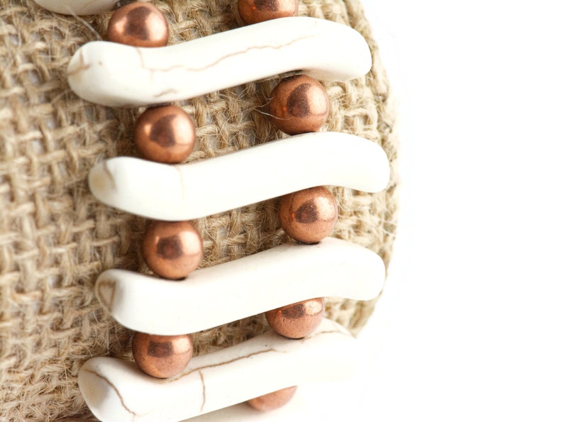 May include: A close-up of a bracelet made with white ceramic beads and copper-colored metal beads. The bracelet is wrapped around a piece of burlap fabric.