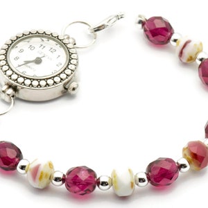 K&ouml;nnte beinhalten: Eine silberne Uhr mit wei&szlig;em Zifferblatt und schwarzen Zahlen ist an einem Perlenarmband befestigt. Das Armband besteht aus abwechselnd rosa und wei&szlig;en Perlen.
