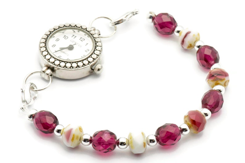 K&ouml;nnte beinhalten: Eine silberne Uhr mit wei&szlig;em Zifferblatt und schwarzen Zahlen ist an einem Perlenarmband befestigt. Das Armband besteht aus abwechselnd rosa und wei&szlig;en Perlen.