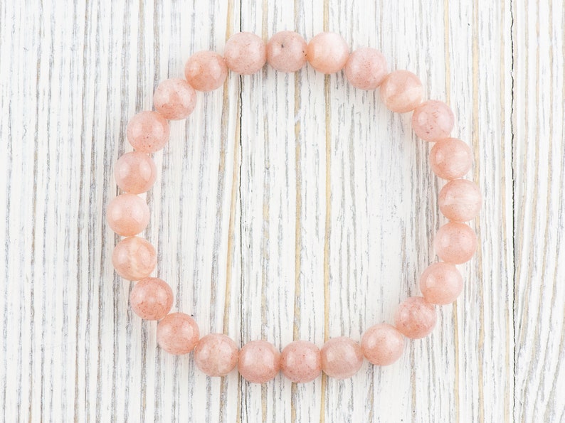 May include: A pink sunstone bracelet with round beads on a white wooden background.