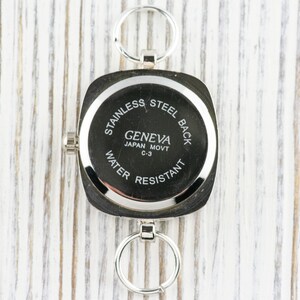 K&ouml;nnte beinhalten: Silberne quadratische Uhr mit einem Edelstahlboden. Das Zifferblatt der Uhr tr&auml;gt den Text "Geneva Japan Movt C-3 Water Resistant".