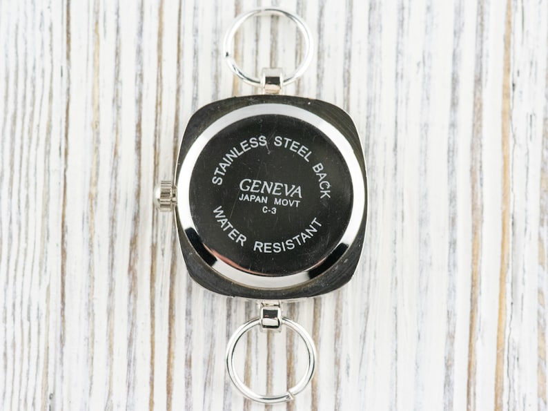 K&ouml;nnte beinhalten: Silberne quadratische Uhr mit einem Edelstahlboden. Das Zifferblatt der Uhr tr&auml;gt den Text "Geneva Japan Movt C-3 Water Resistant".
