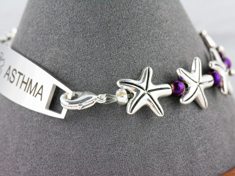 May include: Silver medical ID bracelet with a purple bead and star charm. The bracelet has the word "ASTHMA" engraved on the tag.