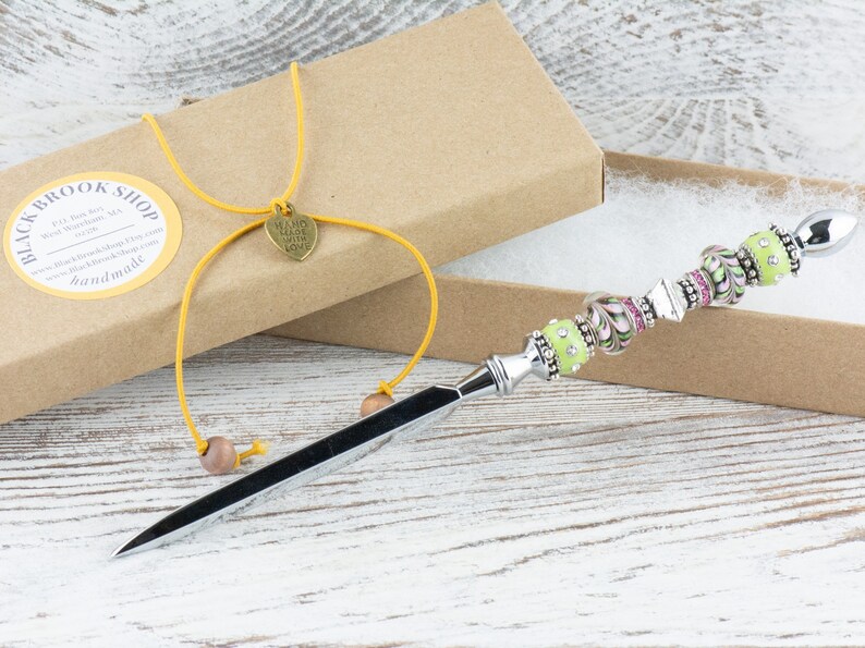 Peut inclure: Un ouvre-lettre en argent avec un manche en perles vertes et roses. Le manche est d&eacute;cor&eacute; de strass et de volutes. L'ouvre-lettre est dans une bo&icirc;te brune avec une ficelle jaune et un charme en forme de c&oelig;ur qui dit "Fait &agrave; la main avec amour".