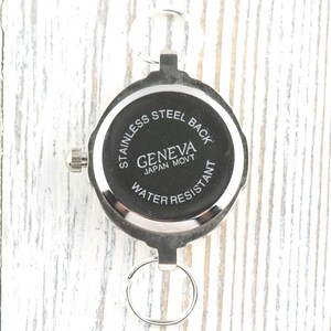 Pu&ograve; includere: Un orologio da tasca argentato con quadrante nero. L'orologio ha un fondello in acciaio inossidabile e recita "GENEVA JAPAN MOVT WATER RESISTANT". L'orologio &egrave; attaccato a un portachiavi.