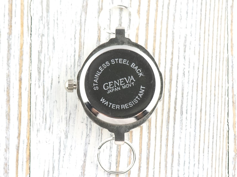 Pu&ograve; includere: Un orologio da tasca argentato con quadrante nero. L'orologio ha un fondello in acciaio inossidabile e recita "GENEVA JAPAN MOVT WATER RESISTANT". L'orologio &egrave; attaccato a un portachiavi.