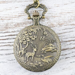 Pu&ograve; includere: Un orologio da tasca in bronzo con un design elaborato che presenta un cervo e una gru in un ambiente boschivo.