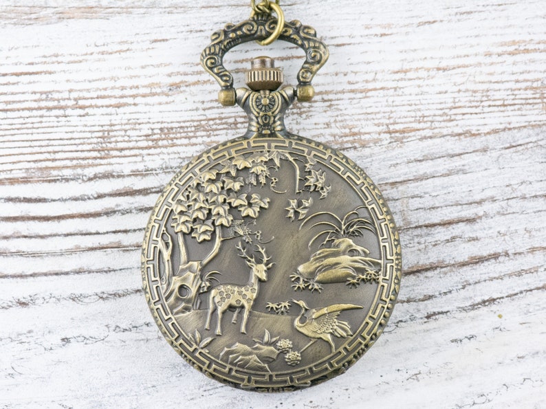 Pu&ograve; includere: Un orologio da tasca in bronzo con un design elaborato che presenta un cervo e una gru in un ambiente boschivo.