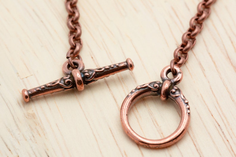Puede incluir: Primer plano de una cadena de cobre con un cierre de palanca adornado. El cierre tiene un dise&ntilde;o decorativo y est&aacute; hecho de una sola pieza de metal.