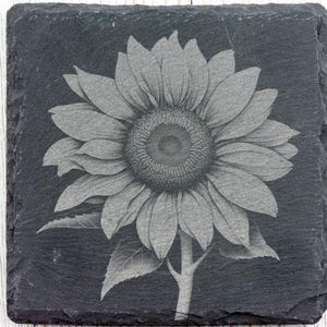 K&ouml;nnte beinhalten: Ein schwarzer Schieferuntersetzer mit einem lasergravierten Design einer grauen Sonnenblume.