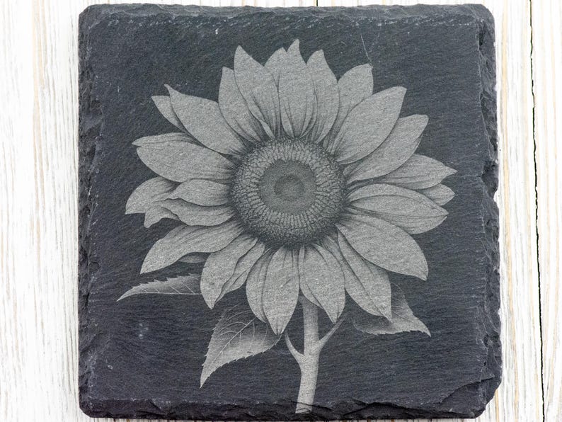 K&ouml;nnte beinhalten: Ein schwarzer Schieferuntersetzer mit einem lasergravierten Design einer grauen Sonnenblume.