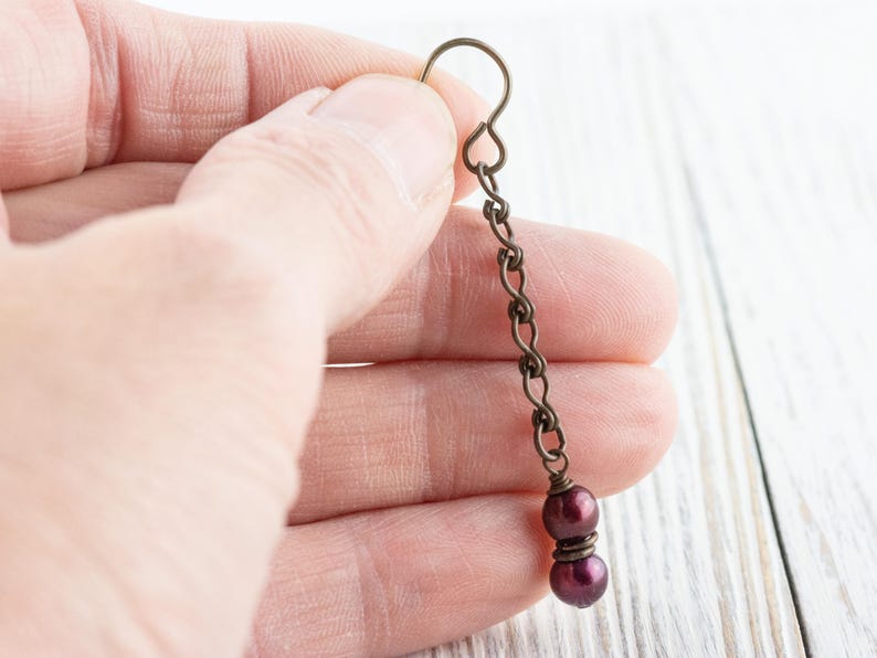 Puede incluir: Un solo pendiente con una cadena y un gancho de bronce oscuro. La cadena suspende dos cuentas de color p&uacute;rpura oscuro. El pendiente se sostiene en la mano de una persona contra un fondo de madera blanca. El pendiente es de estilo colgante.