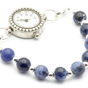 K&ouml;nnte beinhalten: Eine silberne Uhr mit einem Armband aus blauen Edelsteinen. Das Zifferblatt der Uhr hat schwarze r&ouml;mische Ziffern und einen silbernen Sekundenzeiger. Das Armband besteht aus runden blauen Steinen mit silbernen Zwischenst&uuml;cken.