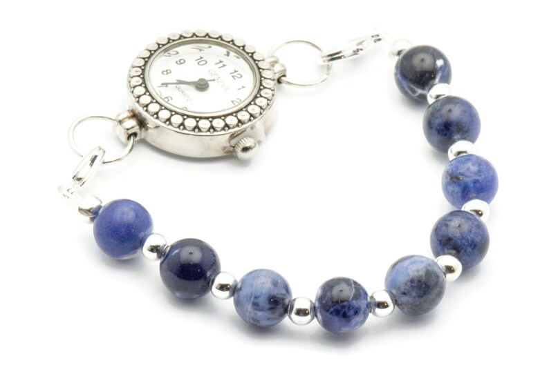 K&ouml;nnte beinhalten: Eine silberne Uhr mit einem Armband aus blauen Edelsteinen. Das Zifferblatt der Uhr hat schwarze r&ouml;mische Ziffern und einen silbernen Sekundenzeiger. Das Armband besteht aus runden blauen Steinen mit silbernen Zwischenst&uuml;cken.
