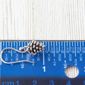 May include: A silver pine cone earring with a hook closure. The earring is measured against a ruler, showing it is just over 1/2 inch long.