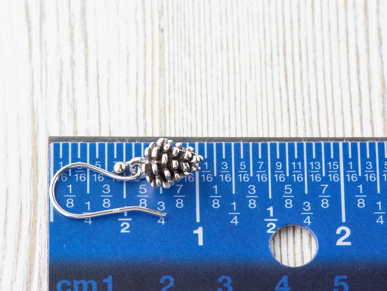 May include: A silver pine cone earring with a hook closure. The earring is measured against a ruler, showing it is just over 1/2 inch long.