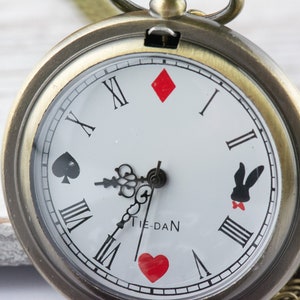 Op de afbeelding: Een vintage-stijl zakhorloge met een witte wijzerplaat en zwarte Romeinse cijfers. Het horloge heeft een vergulde kast en een ketting. De wijzerplaat van het horloge heeft speelkaartsymbolen en een konijnen-silhouet.
